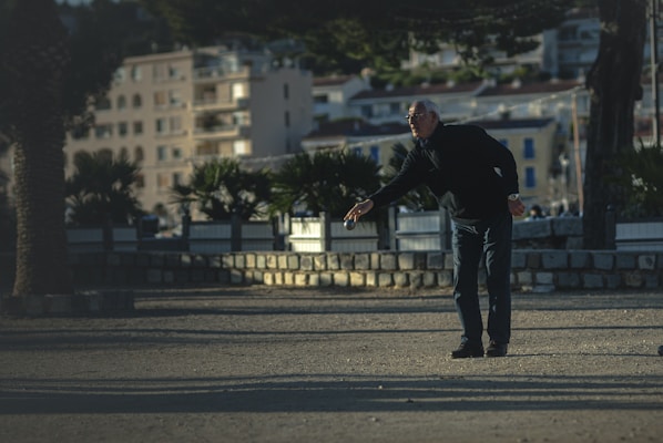 An older man wearing dark clothing is playing a game of pétanque in an open outdoor space surrounded by trees and residential buildings. He appears to be focused as he throws a metal ball, with sunlight casting long shadows on the ground.