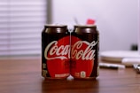 Two cans of Coca-Cola Zero Sugar are placed on a wooden table with a blurred background. The cans are predominantly black with a red circle highlighting the Coca-Cola logo. The setting appears to be indoors with a soft, diffused lighting.