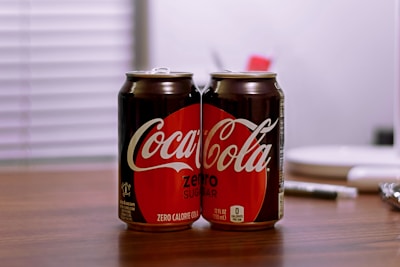 Two cans of Coca-Cola Zero Sugar are placed on a wooden table with a blurred background. The cans are predominantly black with a red circle highlighting the Coca-Cola logo. The setting appears to be indoors with a soft, diffused lighting.