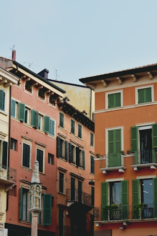 Colorful historical buildings with green shutters and balconies create a quaint and charming European cityscape. The architecture features vibrant orange and terracotta hues and ornate detailing. A small stone monument or column is visible in the foreground, adding to the historical atmosphere.