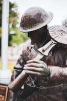 A bronze statue of a soldier wearing a helmet, depicted in a thoughtful pose as he holds onto a gun with a large, circular magazine. The background is slightly blurred, indicating an outdoor setting with trees.