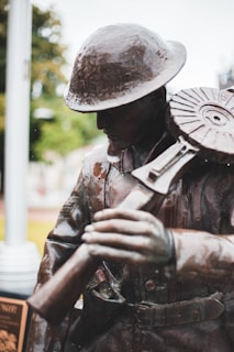 A bronze statue of a soldier wearing a helmet, depicted in a thoughtful pose as he holds onto a gun with a large, circular magazine. The background is slightly blurred, indicating an outdoor setting with trees.