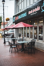 Outdoor café seating with several black chairs and tables arranged on a red brick sidewalk. Red umbrellas provide shade above the tables. The café is adjacent to a building with signage for 'Cycle Solutions' and 'Brewed Awakening Café & Roasting Co.' A hanging flower basket and a streetlamp are visible along the sidewalk.