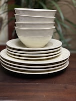 A neatly stacked collection of ceramic bowls and plates, with six bowls positioned on top of four plates. The ceramics have a clean, minimalist design with subtle off-white hues, against a wooden surface. Faint greenery is blurred in the background.