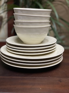 A neatly stacked collection of ceramic bowls and plates, with six bowls positioned on top of four plates. The ceramics have a clean, minimalist design with subtle off-white hues, against a wooden surface. Faint greenery is blurred in the background.