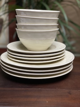 A neatly stacked collection of ceramic bowls and plates, with six bowls positioned on top of four plates. The ceramics have a clean, minimalist design with subtle off-white hues, against a wooden surface. Faint greenery is blurred in the background.