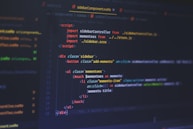 A computer screen displays code written in a text editor. The code is related to web development and includes HTML and JavaScript. The interface shows a dark theme with syntax highlighting, featuring different colors for commands, variables, and elements. Some parts of the screen show filenames and directories on the left.