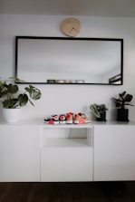 A modern, minimalist interior setup with a white cabinet, decorated with three small pairs of shoes placed on top. A large rectangular mirror with a black frame hangs above the cabinet, and a uniquely shaped wall clock is positioned above the mirror. Two potted plants are placed on either side of the cabinet, adding a touch of greenery.
