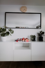 A modern, minimalist interior setup with a white cabinet, decorated with three small pairs of shoes placed on top. A large rectangular mirror with a black frame hangs above the cabinet, and a uniquely shaped wall clock is positioned above the mirror. Two potted plants are placed on either side of the cabinet, adding a touch of greenery.
