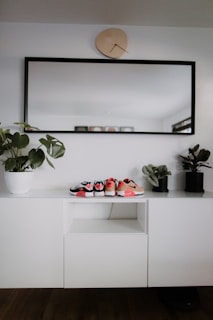 A modern, minimalist interior setup with a white cabinet, decorated with three small pairs of shoes placed on top. A large rectangular mirror with a black frame hangs above the cabinet, and a uniquely shaped wall clock is positioned above the mirror. Two potted plants are placed on either side of the cabinet, adding a touch of greenery.