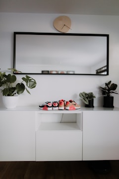 A modern, minimalist interior setup with a white cabinet, decorated with three small pairs of shoes placed on top. A large rectangular mirror with a black frame hangs above the cabinet, and a uniquely shaped wall clock is positioned above the mirror. Two potted plants are placed on either side of the cabinet, adding a touch of greenery.