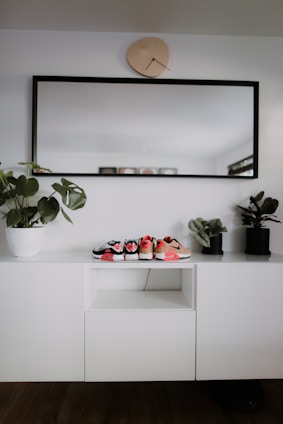 A modern, minimalist interior setup with a white cabinet, decorated with three small pairs of shoes placed on top. A large rectangular mirror with a black frame hangs above the cabinet, and a uniquely shaped wall clock is positioned above the mirror. Two potted plants are placed on either side of the cabinet, adding a touch of greenery.
