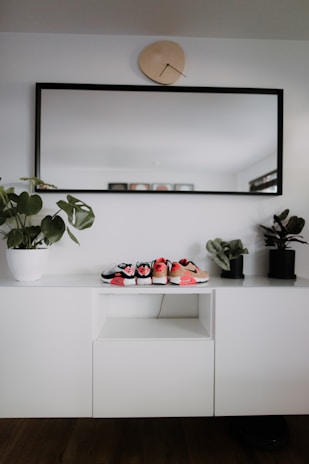 A modern, minimalist interior setup with a white cabinet, decorated with three small pairs of shoes placed on top. A large rectangular mirror with a black frame hangs above the cabinet, and a uniquely shaped wall clock is positioned above the mirror. Two potted plants are placed on either side of the cabinet, adding a touch of greenery.