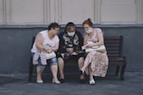 Three elderly women are sitting on a bench outside, engaging with something in the hands of the middle woman. They are wearing masks and appear to be focused and engaged. The setting looks urban, with a gray wall and pavement.