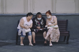 Three elderly women are sitting on a bench outside, engaging with something in the hands of the middle woman. They are wearing masks and appear to be focused and engaged. The setting looks urban, with a gray wall and pavement.