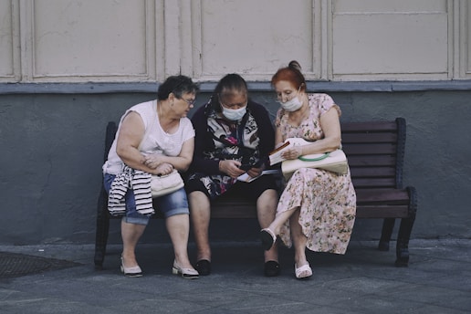 Three elderly women are sitting on a bench outside, engaging with something in the hands of the middle woman. They are wearing masks and appear to be focused and engaged. The setting looks urban, with a gray wall and pavement.