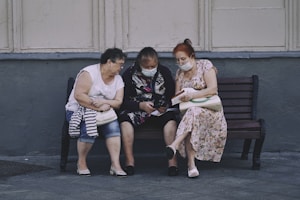 Three elderly women are sitting on a bench outside, engaging with something in the hands of the middle woman. They are wearing masks and appear to be focused and engaged. The setting looks urban, with a gray wall and pavement.