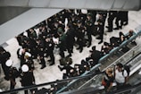 A group of uniformed individuals, possibly military personnel, are gathered in an indoor setting, standing and sitting in an orderly manner. Some are wearing white caps, while most have dark uniforms. Two civilians are seen on an escalator to the right.
