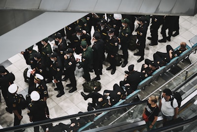 A group of uniformed individuals, possibly military personnel, are gathered in an indoor setting, standing and sitting in an orderly manner. Some are wearing white caps, while most have dark uniforms. Two civilians are seen on an escalator to the right.