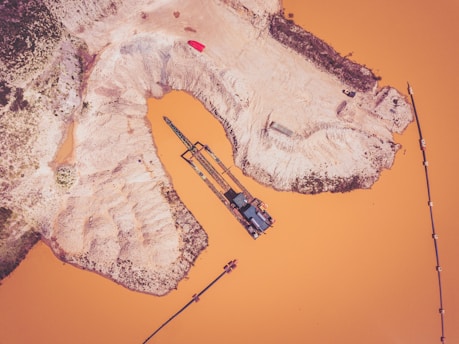 An aerial view of an industrial area with a large body of orange-colored water surrounded by dirt and sand. There is a long, segmented structure extending into the water, which appears to be a conveyor belt or some kind of industrial rig. The landscape around the water is barren and rugged, with some vegetation visible at the upper left edge.