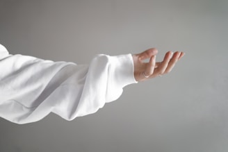 A hand extends outward with an open palm, partially covered by a white sleeve against a gray background. The fingers are slightly curled in a relaxed and inviting gesture.