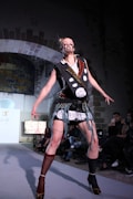 A model on a runway is dressed in a bold, avant-garde outfit. The attire involves a lot of black and white elements with intricate patterns and patchwork. The face is marked with distinct makeup, adding a dramatic effect. The background shows an audience seated in a dimly lit room with arched architecture.