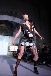 A model on a runway is dressed in a bold, avant-garde outfit. The attire involves a lot of black and white elements with intricate patterns and patchwork. The face is marked with distinct makeup, adding a dramatic effect. The background shows an audience seated in a dimly lit room with arched architecture.