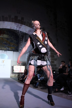 A model on a runway is dressed in a bold, avant-garde outfit. The attire involves a lot of black and white elements with intricate patterns and patchwork. The face is marked with distinct makeup, adding a dramatic effect. The background shows an audience seated in a dimly lit room with arched architecture.