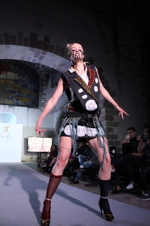 A model on a runway is dressed in a bold, avant-garde outfit. The attire involves a lot of black and white elements with intricate patterns and patchwork. The face is marked with distinct makeup, adding a dramatic effect. The background shows an audience seated in a dimly lit room with arched architecture.