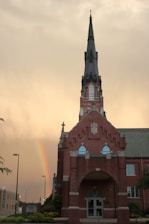 A tall brick church with a prominent steeple is set against a dramatic sky. A vibrant rainbow is visible in the backdrop, enhancing the serene and hopeful atmosphere. The building's architecture features Gothic elements, including pointed arches and decorative stonework.