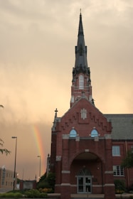 A tall brick church with a prominent steeple is set against a dramatic sky. A vibrant rainbow is visible in the backdrop, enhancing the serene and hopeful atmosphere. The building's architecture features Gothic elements, including pointed arches and decorative stonework.
