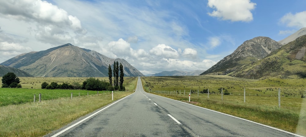 camino de concreto gris cerca del campo de hierba verde y la montaña bajo nubes blancas y cielo azul durante