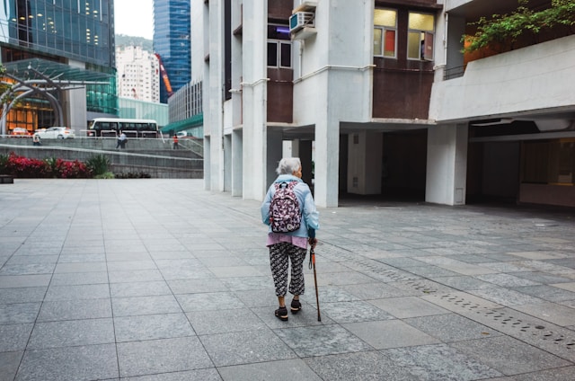 An older person walking with a cane in an urban environment. The individual is wearing patterned pants, a top, and a backpack. Surrounding them are modern buildings with glass facades and a bus is visible in the distance.