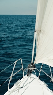 The image captures the bow of a sailboat navigating through the ocean with part of a white sail visible. The sea appears vast and slightly turbulent, with small waves and a deep blue color. The horizon is faint and the sky is clear, suggesting a calm weather.