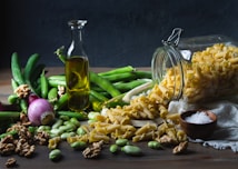 A variety of fresh ingredients are scattered on a wooden surface. A glass bottle of olive oil stands upright, surrounded by green vegetables, including cucumbers and beans. A red onion is present on the left side, while a glass jar is tipped over, spilling pasta. Walnuts and broad beans are also spread out alongside a small wooden bowl filled with coarse salt.