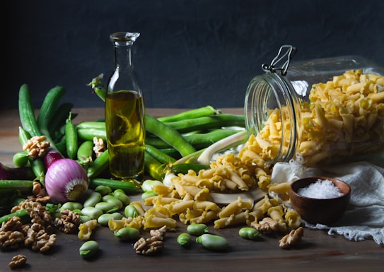 A variety of fresh ingredients are scattered on a wooden surface. A glass bottle of olive oil stands upright, surrounded by green vegetables, including cucumbers and beans. A red onion is present on the left side, while a glass jar is tipped over, spilling pasta. Walnuts and broad beans are also spread out alongside a small wooden bowl filled with coarse salt.