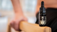 A small black bottle labeled 'Hemp Crew Bourbon Vanilla' sits on a wooden surface. The focus is on the bottle, with a hand and blurred background visible, suggesting a casual setting.