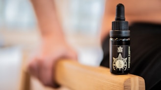 A small black bottle labeled 'Hemp Crew Bourbon Vanilla' sits on a wooden surface. The focus is on the bottle, with a hand and blurred background visible, suggesting a casual setting.