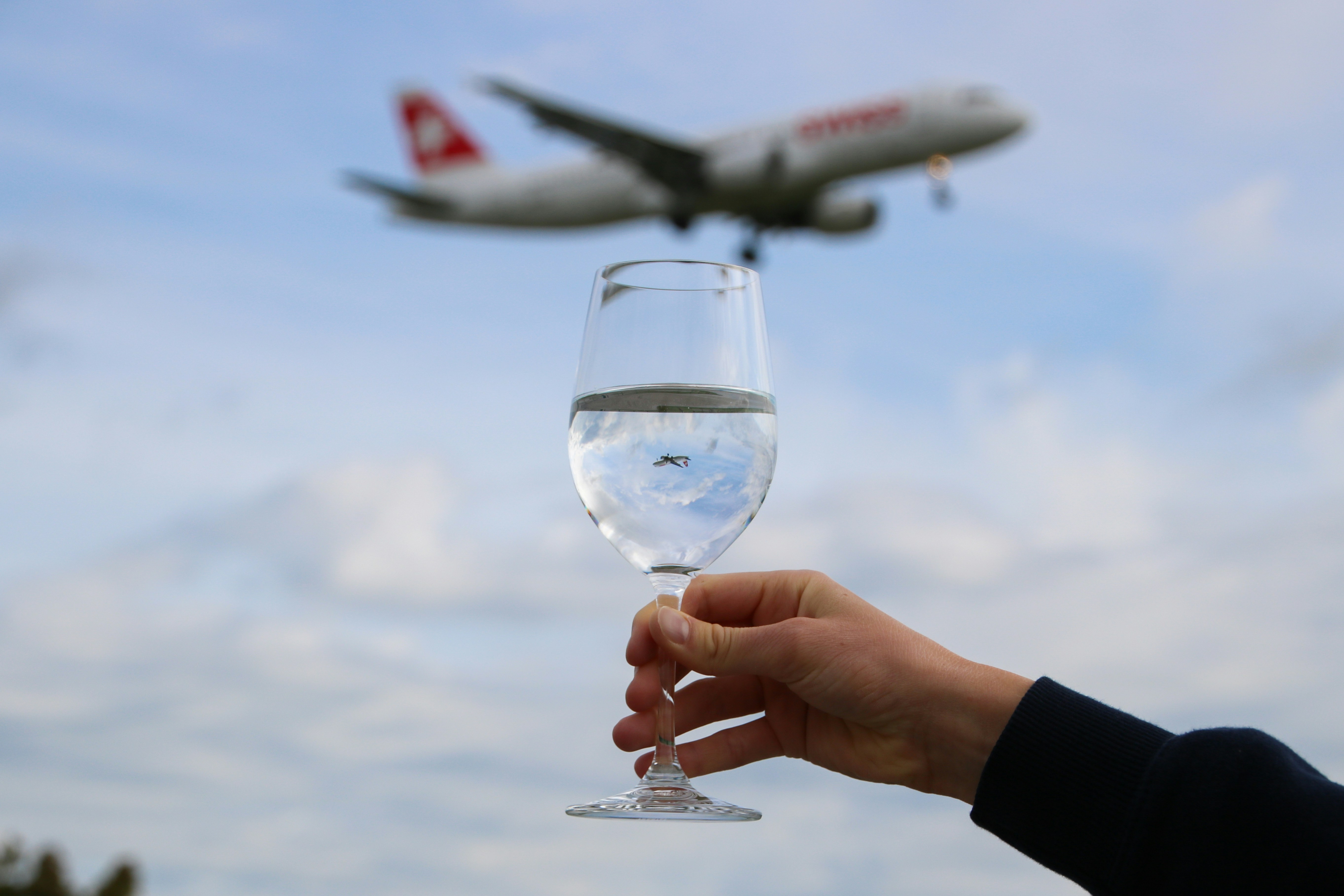 person holding clear wine glass with white airplane in the sky, a landing swiss international airlines plane reflecting in a water glass at zurich airport 