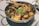 A metal pan contains a mix of seafood including shrimp and crab, garnished with chopped green onions. Corn pieces are also visible among the seafood which seems to be cooked with a spicy sauce.