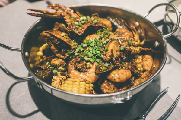 A metal pan contains a mix of seafood including shrimp and crab, garnished with chopped green onions. Corn pieces are also visible among the seafood which seems to be cooked with a spicy sauce.
