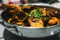 A close-up view of a metal dish filled with cooked seafood, possibly consisting of crabs or lobsters, garnished with chopped green onions. The seafood appears to be seasoned and cooked in a rich, savory sauce, with some corn evident in the dish. The presentation suggests a warm and enticing meal.
