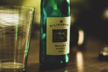 A wine bottle labeled 'Kilikanoon Mulga Shiraz 2015' stands on a wooden surface next to a transparent glass with a decorative pattern. The scene is softly lit with a warm ambiance, creating a cozy and intimate atmosphere.