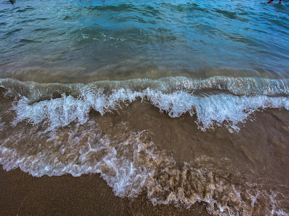 The image depicts a serene beach scene with light brown sand and deep blue water. The waves are crashing onto the shore, creating a rhythmic pattern that adds to the overall tranquility of the setting. The perspective is from above, providing an expansive view of the ocean meeting the sky at the horizon line.