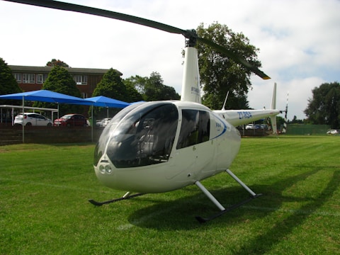A white helicopter with registration markings sits on a grassy area. Behind the helicopter, there are several parked cars and a building with trees surrounding the area. The sky is partly cloudy.