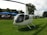 A white helicopter with registration markings sits on a grassy area. Behind the helicopter, there are several parked cars and a building with trees surrounding the area. The sky is partly cloudy.
