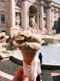 A hand holding a cone of pistachio gelato with a historic fountain in the background featuring classical sculptures and architecture.