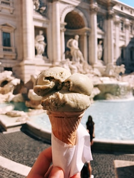 A hand holding a cone of pistachio gelato with a historic fountain in the background featuring classical sculptures and architecture.