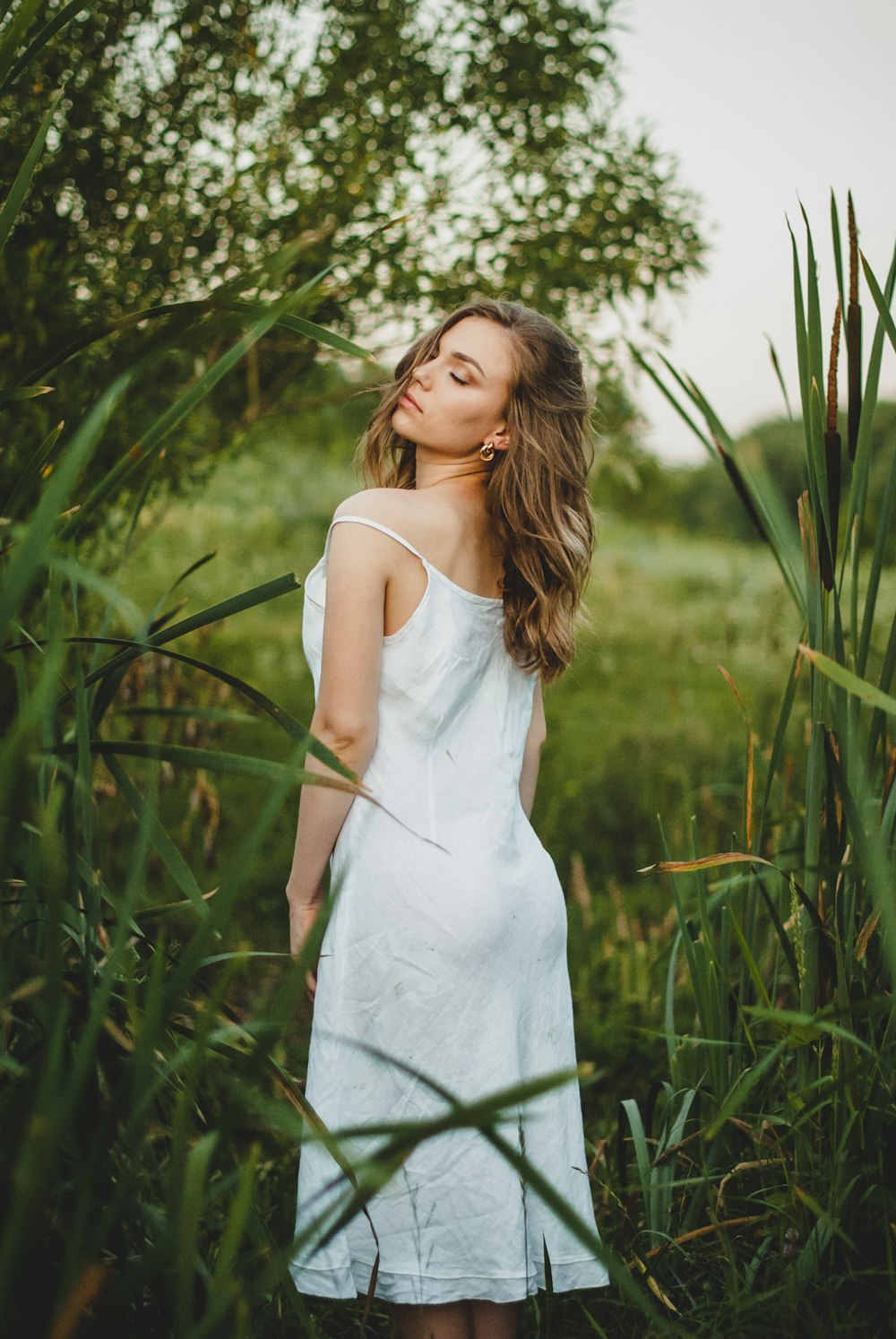 낮 동안 녹색 식물 근처에 서 있는 흰색 스파게티 스트랩 드레스를 입은 여자