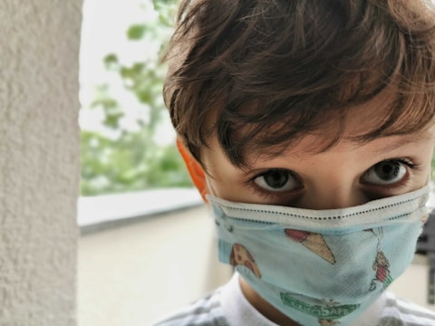 A young child with wavy hair is wearing a face mask decorated with colorful images of ice cream and pizza. The large, expressive eyes are focused directly towards the viewer. In the background, there is a blurry view of greenery, suggesting an outdoor setting.